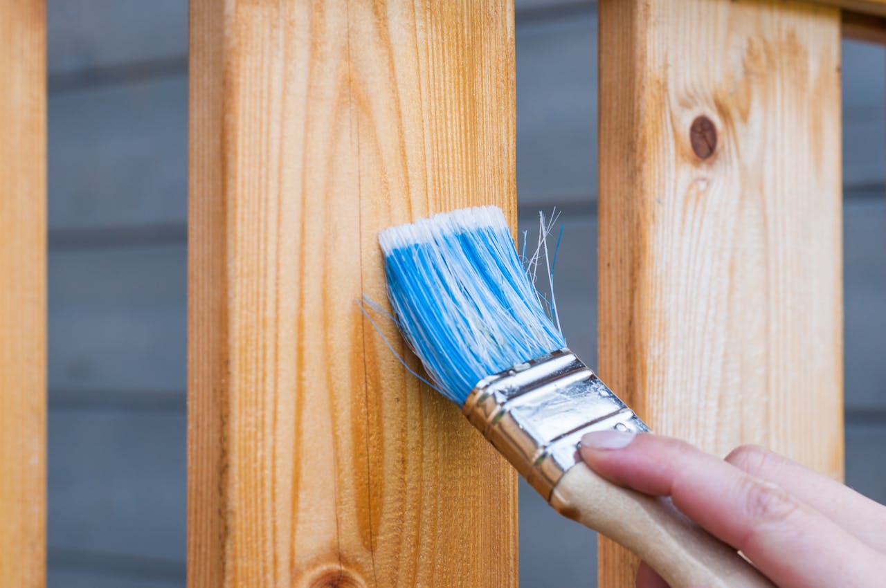 about-img A hand painting a wooden fence plank with a blue paintbrush outdoors. Perfect for DIY and renovation themes.