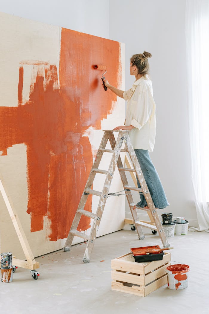 gallery-6 A woman stands on a stepladder, painting a room's wall during home renovation.